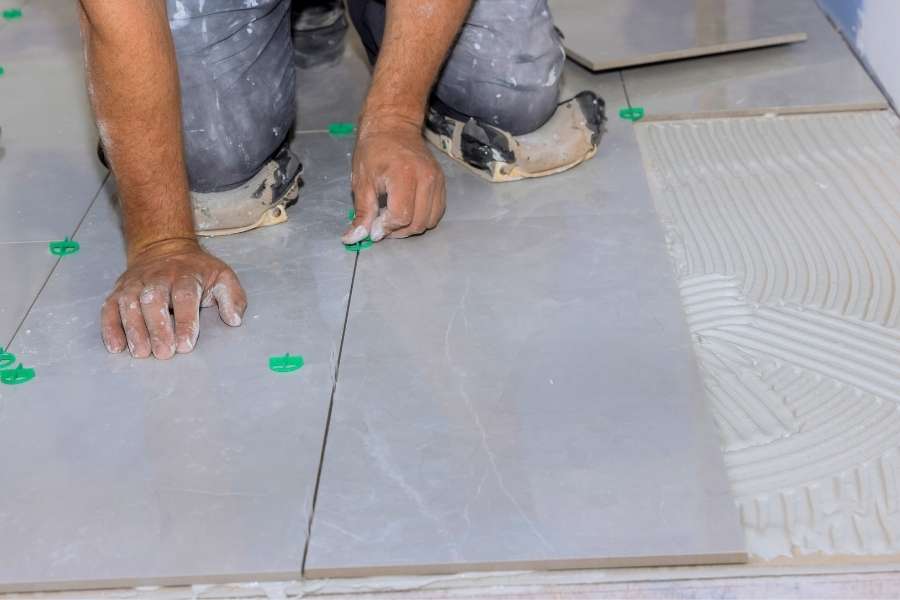 Un carreleur pose du carrelage gris sur un sol en utilisant des croisillons verts pour un alignement parfait.
