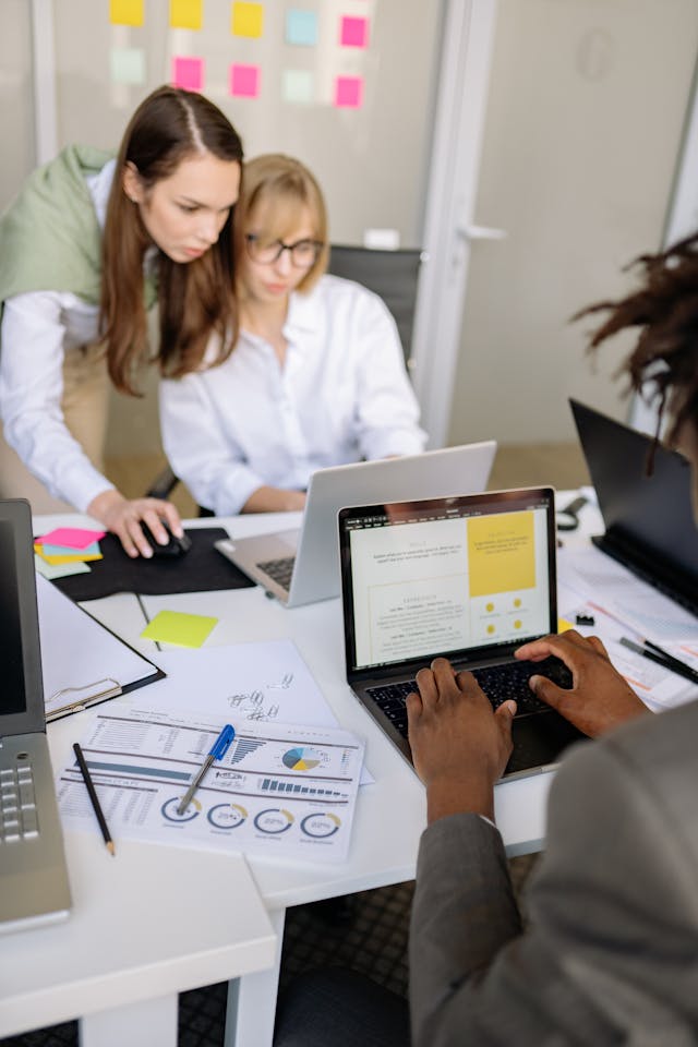 Deux collègues de travail debout se penchent sur l'écran d'un ordinateur portable tandis qu'un homme tape sur son clavier au premier plan, des rapports avec des graphiques sont posés sur la table, et des post-its sont collés au mur.