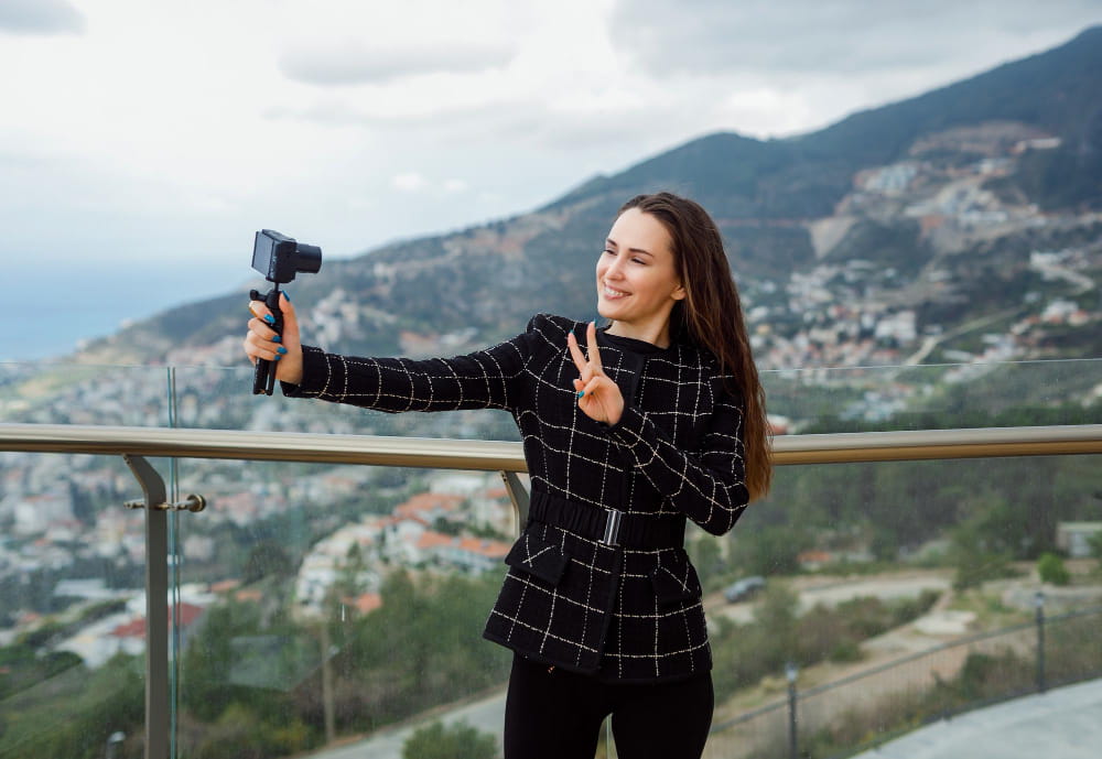 Une jeune femme souriante filme un vlog sur un balcon face à un paysage montagneux, illustrant l'action sans limites grâce à la caméra d'action ultime qu'elle tient sur un trépied portable.