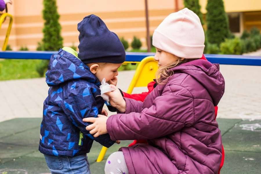 Une grande sœur aide son petit frère à se nettoyer le visage dans un parc, illustrant la complexité de la relation et de la jalousie entre frères et sœurs.